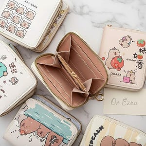 May include: A collection of white and pastel-coloured zip-around wallets, each adorned with cartoon bear illustrations. One wallet is open, displaying a pink interior with a zipped coin pocket. The wallets feature diverse designs and text, including "APY BARA" and "KEEP CALM".