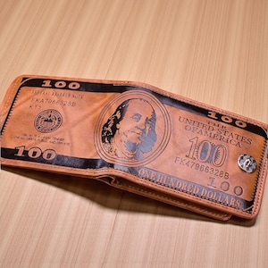 May include: Brown bi-fold wallet designed to look like a $100 bill. The wallet has a black border and includes text such as "FEDERAL RESERVE NOTE" and "ONE HUNDRED DOLLARS". A silver snap closure is visible.