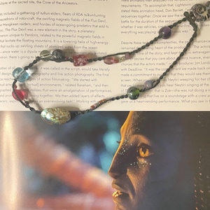 Puede incluir: Un collar de cuentas de colores, incluyendo azules, rosas y verdes, con detalles plateados. Presenta una cuenta ovalada de concha de abulón. El collar se muestra sobre una página impresa con texto.