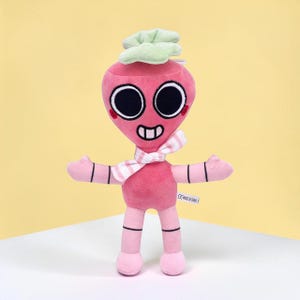 May include: A pink plush toy in the shape of a radish with a smiling face, large black eyes, and a striped scarf. The toy has outstretched arms and a green leafy top. The toy is standing on a white surface against a yellow background.