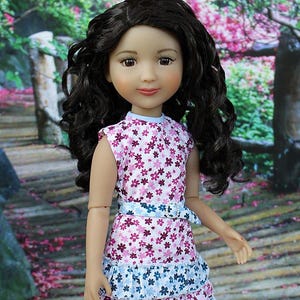 Puede incluir: Una muñeca con el pelo largo, oscuro y rizado que lleva un vestido floral blanco y rosa con un ribete de volantes azules. La muñeca tiene ojos marrones y lleva un top azul claro sin mangas debajo del vestido.