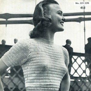 May include: A black and white photograph of a woman wearing a white knitted jumper with short sleeves. The jumper has a textured knit pattern. The woman is smiling and looking to the right. The photo is labelled "Jaeger" and has the number "3211/3298" printed on it.
