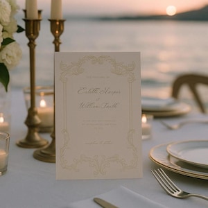 Può includere: Un invito di nozze color crema con bordi decorativi dorati e testo, inclusi i nomi Estelle Harper e William Smith. L'invito è su un tavolo con candelabri dorati e una forchetta.