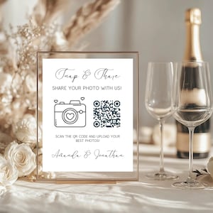 Può includere: Un cartello con cornice dorata con la scritta "Snap & Share" e una grafica a forma di fotocamera, che invita gli ospiti a condividere le foto. Include un codice QR e i nomi "Amanda & Jonathan". Due calici e una bottiglia di champagne sullo sfondo.