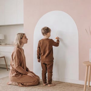 May include: A large, white, arch-shaped wall decal applied to a light pink wall. The decal features a smooth, solid surface, providing a blank space for drawing or writing. It is positioned vertically, extending from the floor upward.