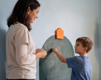 May include: A dark green arched chalkboard mounted on a wall with a small orange pentagonal piece attached to the top. The chalkboard surface shows faint white chalk markings and is positioned vertically against a light blue wall.