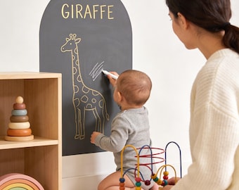 May include: An arched dark gray wall decal featuring the word GIRAFFE in yellow text above a yellow line drawing of a giraffe. The decal is applied to a wall, and a white marker is used to draw on the surface of the dark gray material.
