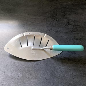 May include: A silver-colored metal kitchen tool with a semi-circular, slotted head and a bright turquoise handle. The metal surface features the embossed text "Toley" and "WELL-DONE" along with "PAT. PEND. PENDING" near the handle attachment point.