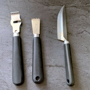 Könnte beinhalten: Drei Küchenutensilien mit schwarzen Griffen: ein Zestenreißer, eine Reibe und ein Messer mit gebogener Klinge. Der Zestenreißer und die Reibe haben Klingen aus Edelstahl. Das Messer hat eine silberne Klinge. Alle drei Werkzeuge dienen der Zubereitung von Speisen.