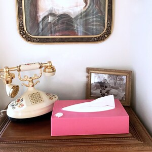 May include: A pink tissue box with a white tissue inside. The tissue box is sitting on a wooden table next to a vintage rotary phone and a framed photo.