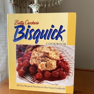 May include: A cookbook titled "Betty Crocker's Bisquick Cookbook" with a yellow cover. The cover features the Bisquick logo in blue and a photo of a cherry cobbler. The text "All Your Bisquick Favorites in One Great Cookbook!" is also visible.