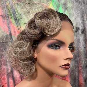 May include: A blonde wig with a vintage style, featuring a curled updo. The wig is styled with a side part and has a lace front.