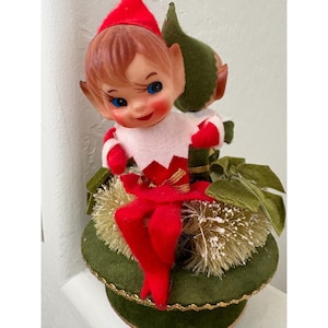 Może przedstawiać: Figurka vintage elfa bożonarodzeniowego z czerwoną czapką, czerwonym strojem i białym kołnierzem. Elf ma różowe policzki i niebieskie oczy. Jest osadzony na zielonej podstawie ze złotym wykończeniem i dekoracyjnymi akcentami.