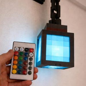 Pode incluir: Uma lâmpada LED preta e cinza em forma de cubo com luz azul, montada em uma parede branca. A lâmpada é controlada por um controle remoto com opções de cores. A lâmpada tem um design geométrico único e é uma luminária moderna.