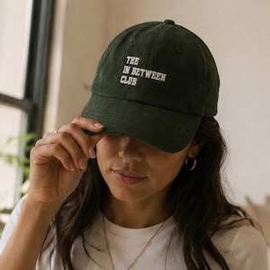 May include: A dark green baseball cap with the text THE IN BETWEEN CLUB embroidered in white on the front. The cap features a structured crown, a curved brim, and a fabric strap closure at the back, providing a classic look for casual wear.