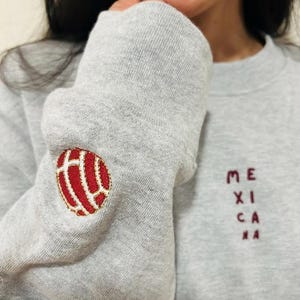 May include: A gray sweatshirt with a red and white embroidered design on the sleeve and the word "MEXICANA" in maroon letters. The sweatshirt is made of a soft material.