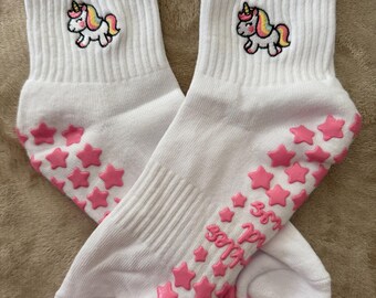 Puede incluir: Un par de calcetines blancos con un unicornio bordado de cuerno amarillo y crin arcoíris. Las suelas tienen agarres de silicona en forma de estrella de color rosa, y el texto "sparkles and smiles" está impreso en rosa en la parte inferior.