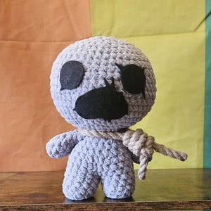 May include: A handmade crochet doll in a light gray color, featuring large black eyes and mouth, and a rope around its neck. The doll is standing on a wooden surface with a colorful background.