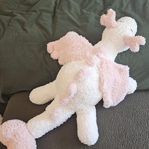 May include: A plush stuffed animal in the shape of a dragon with a white body and soft pink accents on the wings, tail, and head. The creature features a long tail, two wings, and small rounded ears, all crafted from a fuzzy, textured material.