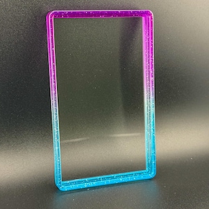 May include: A rectangular, clear frame with a gradient of colors. The top edge is purple, transitioning to pink, then to blue at the bottom. The frame has glitter embedded throughout the edges.