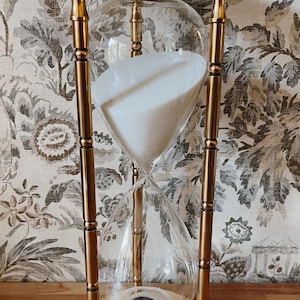 May include: An hourglass with a clear glass bulb filled with white sand, supported by a gold-colored metal frame. The hourglass is set against a floral patterned background. The base and top are circular.