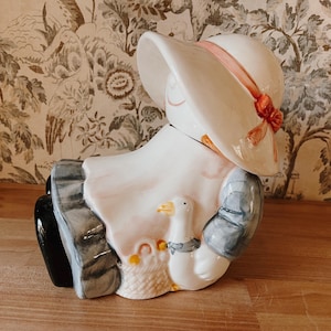 May include: A ceramic cookie jar in the shape of a seated girl wearing a white hat with a pink ribbon. The girl is holding a white goose with a blue scarf and a basket with yellow accents. The jar has a white, pink, blue, and black color scheme.