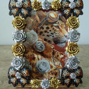 May include: A rectangular picture frame decorated with metallic gold and silver roses along the edges and leopard print bows at the four corners. The frame contains an image of a leopard wearing aviator goggles against a warm-toned background.