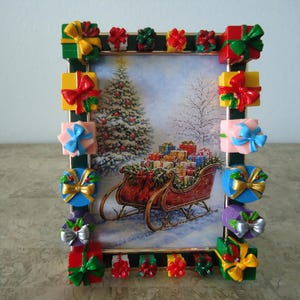 May include: A rectangular picture frame with a dark green border decorated with various colorful miniature gift boxes featuring bows. The center image depicts a red sleigh filled with wrapped presents in front of a snow-covered Christmas tree.