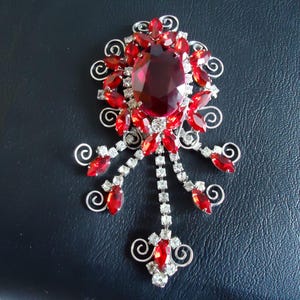 May include: A vintage brooch with a silver-tone metal setting. The brooch features a large, oval, deep red gemstone at its center, surrounded by smaller red and clear crystals. Decorative swirls and dangling crystal accents complete the design.