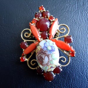 May include: A vintage brooch with a gold-tone filigree base. The brooch features a central oval with a floral design, surrounded by red and orange stones. The brooch has a symmetrical design with a mix of shapes and colors.
