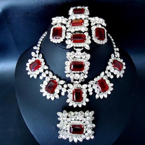 May include: A red and silver rhinestone necklace and bracelet set. The necklace features a large red stone in the center and smaller red stones on either side. The bracelet has a similar design with a smaller red stone in the center.