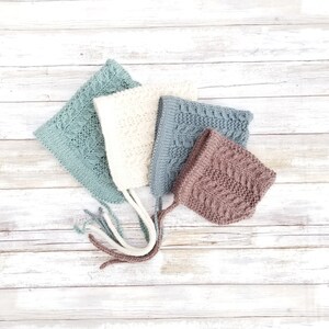 May include: Four knitted baby bonnets in different colors and patterns. The bonnets are made of soft yarn and have a variety of textures. The bonnets are arranged on a white wooden background.