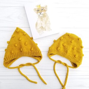 Puede incluir: Dos gorros de bebé de punto amarillo con pompones. Los gorros tienen lazos que se pueden atar debajo de la barbilla.
