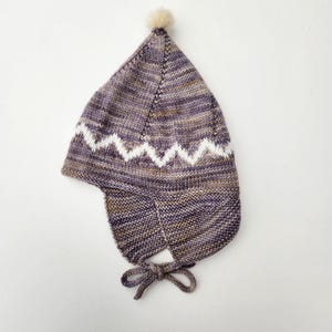 Puede incluir: Un gorro de punto en tonos morados, marrones y blancos, con un patrón en zigzag y orejeras. El gorro tiene un pequeño pompón de color claro en la parte superior y lazos debajo de la barbilla.