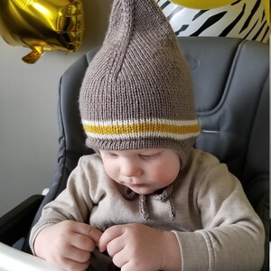 May include: A baby wearing a brown knitted hat with a yellow and white stripe. The hat has a long flap that covers the ears and ties at the chin.