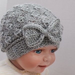 Könnte beinhalten: Eine graue gestrickte Baby-Mütze mit einer großen Schleife oben. Die Mütze ist mit einem strukturierten Stichmuster gearbeitet.