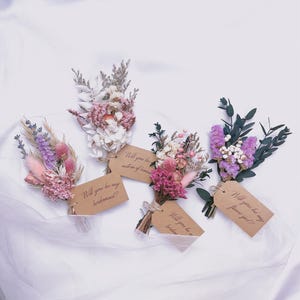Può includere: Cinque piccoli bouquet di fiori secchi nei toni del rosa, viola e bianco, ciascuno legato con spago e un'etichetta marrone. Le etichette recitano: "Will you be my bridesmaid?" o "Will you be my flower girl?"