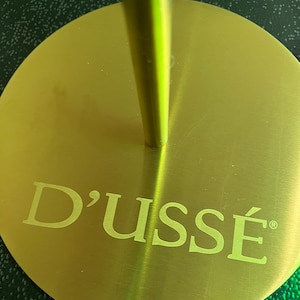 May include: A gold-colored metal stand with the word "D'USSÉ" in a serif font. The stand has a circular base and a vertical stem. The background is a dark green color.