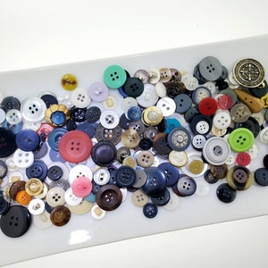 May include: A collection of assorted buttons in various colors, shapes, and sizes. The buttons are made of different materials, including plastic, metal, and wood. Some buttons have a decorative design, while others are plain.