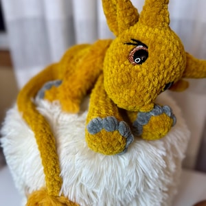May include: A handmade plush toy in the shape of a yellow creature with large ears and gray paws. The toy is made of a soft, textured material and rests on a fluffy white surface. The creature has large, glittery eyes.