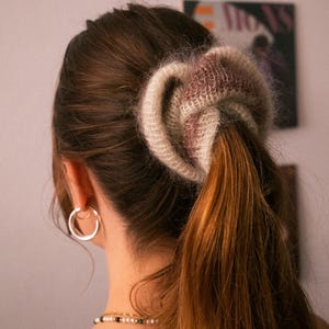 Peut inclure: Gros plan sur une queue de cheval attachée avec une grande chouchou moelleuse. La chouchou est un mélange de tons crème et mauve, complétant les cheveux bruns chauds. Des créoles argentées et un collier délicat sont également visibles.