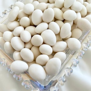 以下が含まれることがあります： 白く楕円形のキャンディーが詰まった透明なガラス皿のクローズアップ。 キャンディーはぎっしりと詰まっており、マットな仕上がりです。 皿にはハート型のアクセントが付いた装飾的な縁があります。