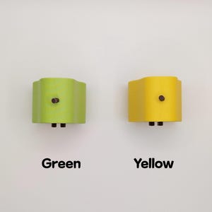 Puede incluir: Dos apliques de pared, uno verde y otro amarillo, est&aacute;n montados en una pared blanca. Cada aplique tiene una parte superior e inferior curvadas, con un bot&oacute;n marr&oacute;n oscuro y dos peque&ntilde;as clavijas negras. Las palabras "Verde" y "Amarillo" est&aacute;n escritas debajo.