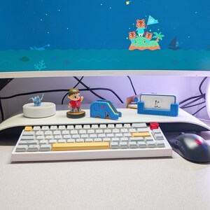 May include: A white mechanical keyboard with yellow and red keycaps sits on a desk. A computer monitor displays a cartoon scene of an island with characters. A mouse and various desk accessories are also visible.
