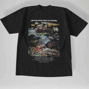 May include: Black t-shirt featuring a vintage Universal Studios Florida graphic. The design includes King Kong, a dinosaur, and the text "SEE THE STARS. RIDE THE MOVIES." The shirt's design is in shades of blue, orange, and yellow.