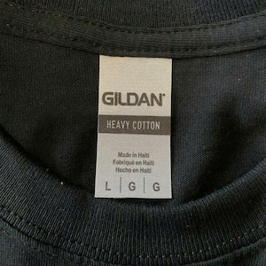 May include: Close-up of a black t-shirt with a tag. The tag reads "GILDAN" at the top, with "HEAVY COTTON" below. The tag also states "Made in Haiti" in English, French, and Spanish. The sizes are L and G.