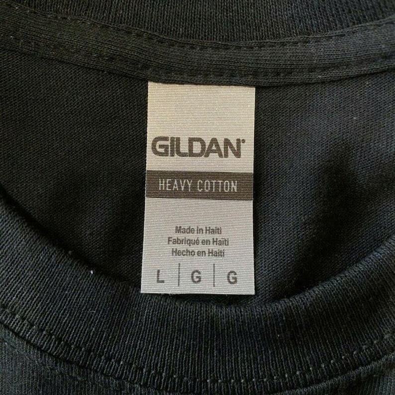 May include: Close-up of a black t-shirt with a tag. The tag reads "GILDAN" at the top, with "HEAVY COTTON" below. The tag also states "Made in Haiti" in English, French, and Spanish. The sizes are L and G.