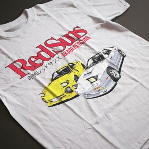 Op de afbeelding: Wit t-shirt met een afbeelding van twee sportwagens, een gele en een witte, en de tekst "RedSuns" in het rood en Japanse karakters. Het shirt is gemaakt van een zachte, lichtgekleurde stof.