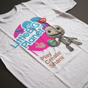 Może przedstawiać: Biała koszulka z logo "Little Big Planet" w kolorze niebieskim i różowym oraz postacią trzymającą globus. Koszulka ma również brązowy napis "Play Create Share".
