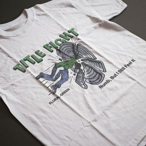 Puede incluir: Camiseta blanca con un dise&ntilde;o gr&aacute;fico. El dise&ntilde;o presenta las palabras "TITLE FIGHT" en letras verdes. La ilustraci&oacute;n muestra a una persona cayendo, con las palabras "FLORAL GREEN" y "Numb, But I Still Feel It" tambi&eacute;n impresas en la camiseta.